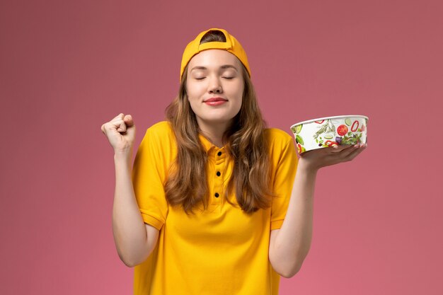 Vue de face femme courrier en uniforme jaune et cape tenant le bol de livraison sur le mur rose service de livraison de travail uniforme de l'entreprise