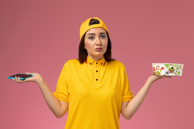 Vue de face femme courrier en uniforme jaune et cape tenant le bol de livraison et smartphone sur la livraison uniforme de service de mur rose clair