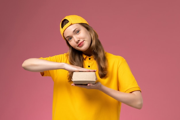 Vue de face femme courrier en uniforme jaune et cape tenant un petit paquet de nourriture de livraison vide sur l'uniforme de livraison de service de mur rose clair