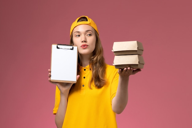 Vue de face femme courrier en uniforme jaune et cape tenant peu de colis alimentaires de livraison avec bloc-notes sur le mur rose service de livraison uniforme fille travailleur