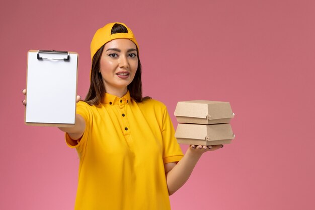 Vue de face femme courrier en uniforme jaune et cape tenant peu de colis alimentaires de livraison avec bloc-notes sur le travail de livraison uniforme de service de mur rose