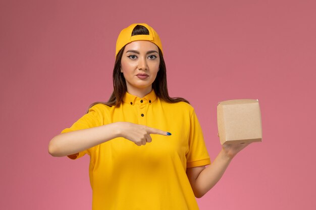 Vue de face femme courrier en uniforme jaune et cape tenant peu de colis alimentaires de livraison sur un mur rose clair entreprise de livraison de services uniformes