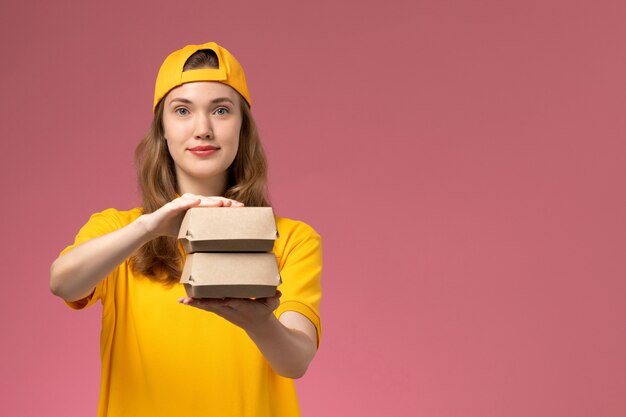 Vue de face femme courrier en uniforme jaune et cape tenant peu de colis alimentaires de livraison sur l'uniforme de livraison des travailleurs de service de mur rose