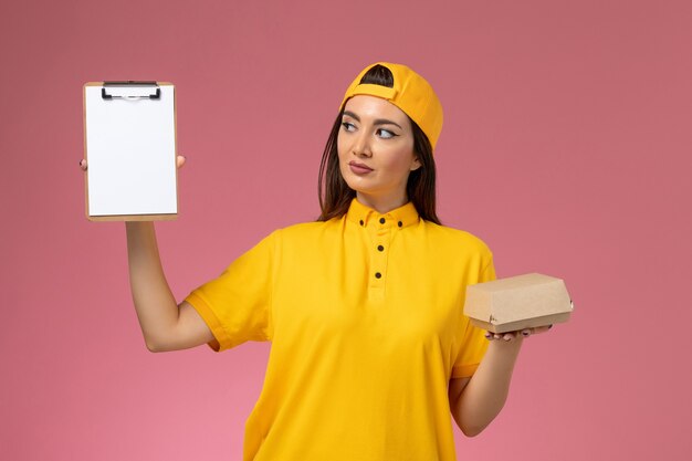 Vue de face femme courrier en uniforme jaune et cape tenant peu de colis de nourriture de livraison et bloc-notes sur mur rose clair service de livraison uniforme entreprise fille travailleur