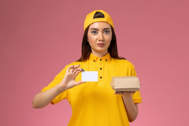 Vue de face femme courrier en uniforme jaune et cape tenant peu de colis de nourriture de livraison et carte sur mur rose clair service de livraison uniforme de travail de travailleur de l'entreprise