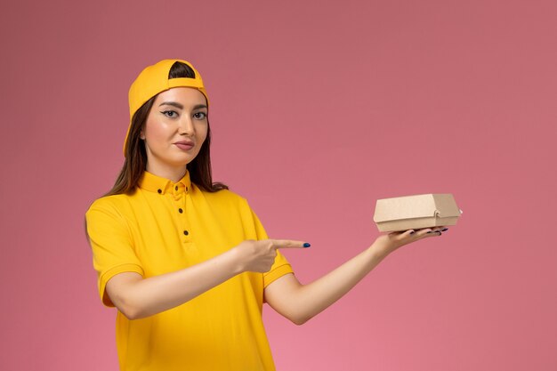 Vue de face femme courrier en uniforme jaune et cape tenant peu de colis de nourriture de livraison sur le mur rose uniforme de livraison de services de l'entreprise de travail des travailleurs