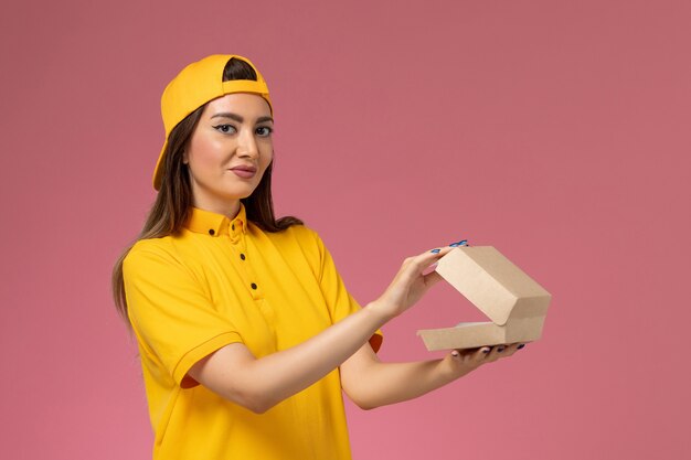 Vue de face femme courrier en uniforme jaune et cape tenant peu de colis de nourriture de livraison et de l'ouvrir sur le mur rose travail de l'entreprise de livraison de services uniforme