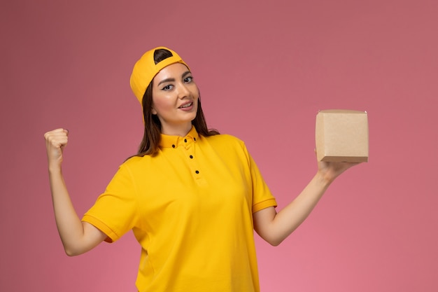 Vue de face femme courrier en uniforme jaune et cape tenant peu de colis de nourriture de livraison se réjouissant sur la société de livraison de services uniformes mur rose clair