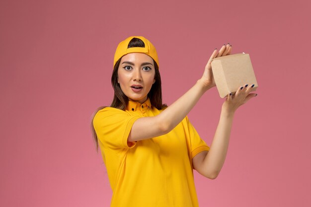 Vue de face femme courrier en uniforme jaune et cape tenant peu de colis de nourriture de livraison sur la société de livraison uniforme de service de mur rose clair