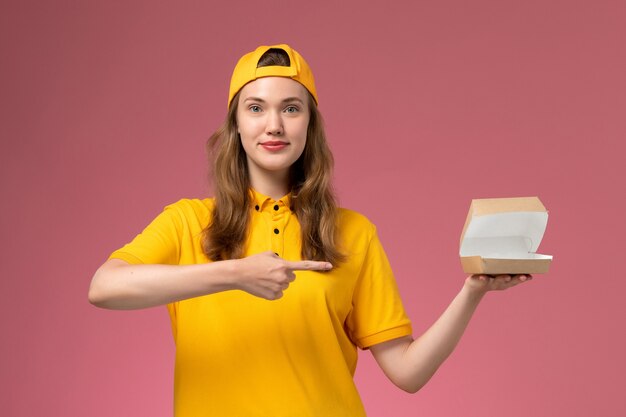 Vue de face femme courrier en uniforme jaune et cape tenant peu d'ouverture de colis de nourriture de livraison sur le mur rose clair travail uniforme de livraison de services