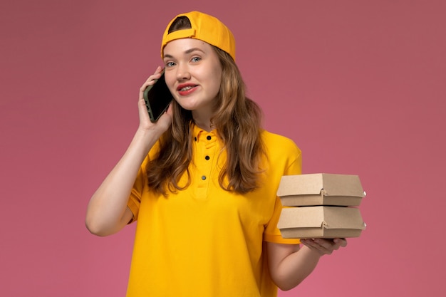 Vue de face femme courrier en uniforme jaune et cape tenant le téléphone avec des paquets de nourriture sur l'uniforme de travail de livraison de services de l'entreprise de mur rose