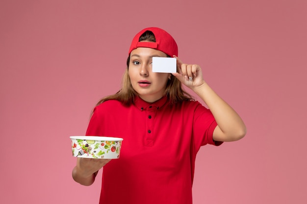 Vue de face femme courrier en uniforme rouge et cape tenant le bol et la carte de livraison sur le mur rose, travailleur de l'emploi de service de livraison uniforme