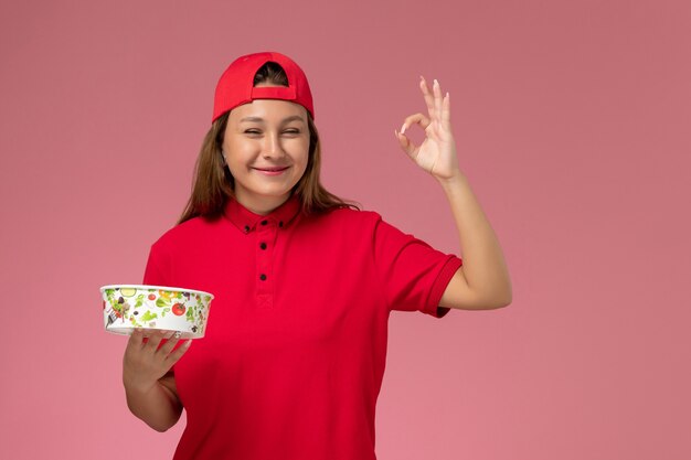 Vue de face femme courrier en uniforme rouge et cape tenant le bol de livraison sur le mur rose, service de livraison uniforme de travailleur d'emploi