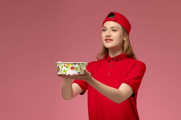 Vue de face femme courrier en uniforme rouge et cape tenant le bol de livraison et souriant sur mur rose clair, service de livraison uniforme