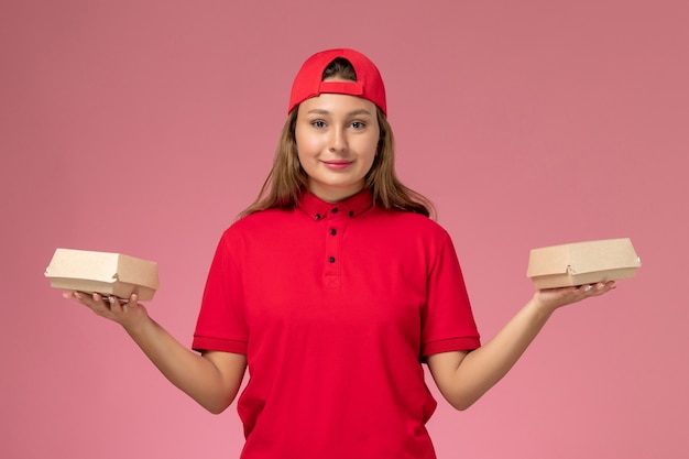 Vue de face femme courrier en uniforme rouge et cape tenant des colis alimentaires de livraison sur un mur rose clair, un service de livraison uniforme entreprise job girl