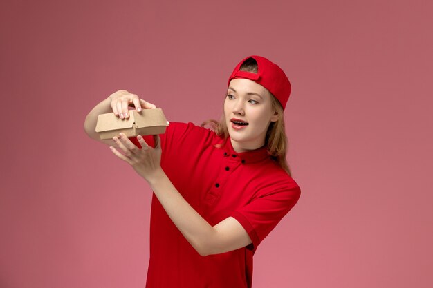 Vue de face femme courrier en uniforme rouge et cape tenant et ouvrant peu de colis de livraison de nourriture sur le mur rose, service de livraison entreprise uniforme