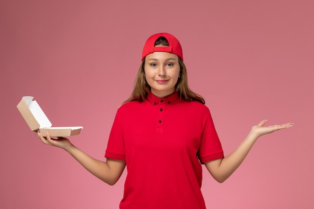 Vue de face femme courrier en uniforme rouge et cape tenant le paquet alimentaire de livraison vide sur mur rose clair, travailleur de l'entreprise de service de livraison uniforme