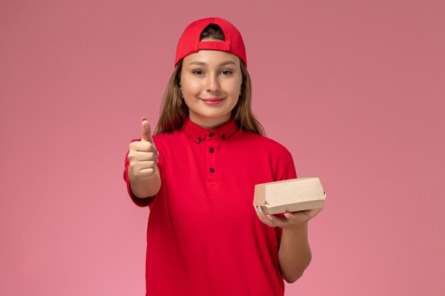 Vue de face femme courrier en uniforme rouge et cape tenant le paquet de nourriture de livraison sur le fond rose service de livraison uniforme travail entreprise
