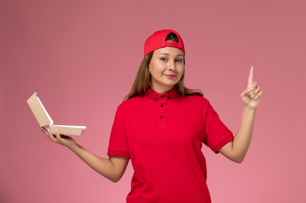 Vue de face femme courrier en uniforme rouge et cape tenant le paquet de nourriture de livraison vide sur mur rose clair, travail de l'entreprise de service de livraison uniforme