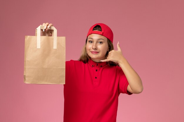 Vue de face femme courrier en uniforme rouge et cape tenant le paquet de papier de livraison sur le mur rose, travail de service de livraison uniforme