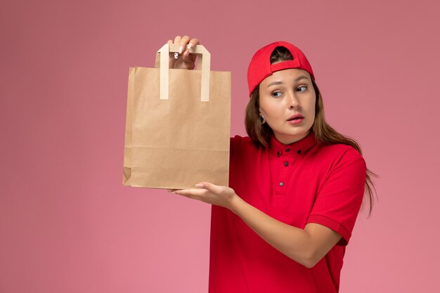 Vue de face femme courrier en uniforme rouge et cape tenant le paquet de papier de livraison sur le mur rose, travailleur de service de livraison uniforme de travail