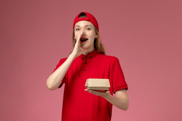 Vue de face femme courrier en uniforme rouge et cape tenant peu de colis alimentaires de livraison appelant sur le mur rose, travail uniforme de l'entreprise de services de livraison