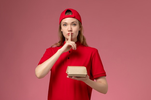 Vue de face femme courrier en uniforme rouge et cape tenant peu de colis alimentaires de livraison sur le mur rose, travail uniforme de l'entreprise de services de livraison