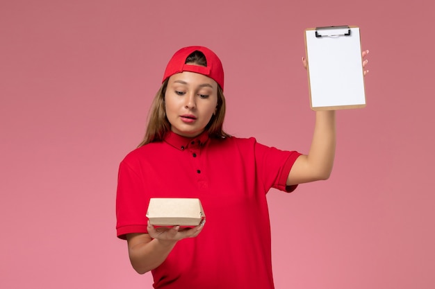 Vue de face femme courrier en uniforme rouge et cape tenant peu de colis de nourriture de livraison et bloc-notes sur mur rose, entreprise de service de livraison uniforme de travailleur