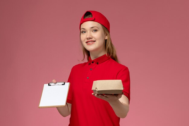 Vue de face femme courrier en uniforme rouge et cape tenant peu de colis de nourriture de livraison avec bloc-notes sur le mur rose, travail de livraison uniforme de service