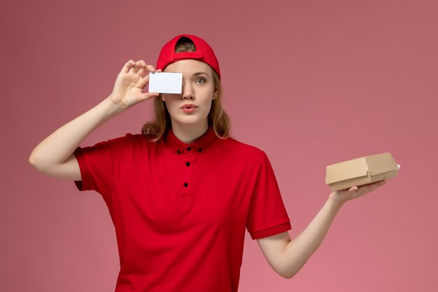 Vue de face femme courrier en uniforme rouge et cape tenant peu de colis de nourriture de livraison avec une carte en plastique blanc sur le mur rose, la livraison d'uniforme de travail de service