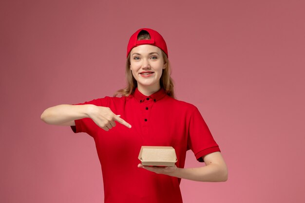 Vue de face femme courrier en uniforme rouge et cape tenant peu de colis de nourriture de livraison souriant sur le mur rose, travail uniforme de l'entreprise de service de livraison