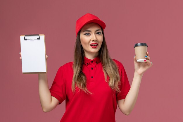 Vue de face femme courrier en uniforme rouge et cape tenant la tasse de café de livraison avec bloc-notes sur fond rose uniforme de travailleur de livraison de services