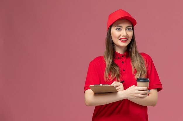 Vue de face femme courrier en uniforme rouge et cape tenant la tasse de café de livraison avec bloc-notes et stylo sur l'uniforme de livraison de service de bureau rose