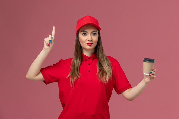 Vue de face femme courrier en uniforme rouge et cape tenant la tasse de café de livraison avec le doigt levé sur l'uniforme de prestation de services fond rose