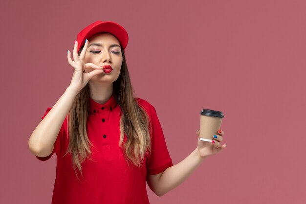 Vue de face femme courrier en uniforme rouge et cape tenant la tasse de café de livraison sur fond rose prestation de services uniforme travail travailleur fille