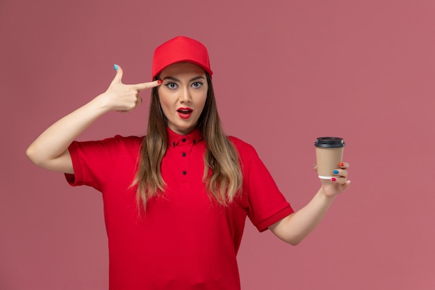 Vue de face femme courrier en uniforme rouge et cape tenant la tasse de café de livraison sur le service de bureau rose travail uniforme de livraison