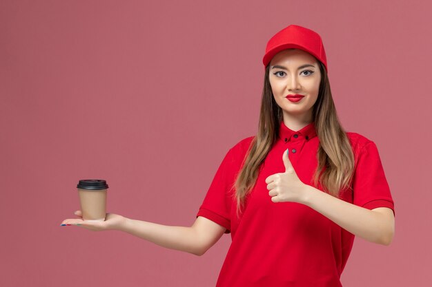 Vue de face femme courrier en uniforme rouge et cape tenant la tasse de café de livraison souriant sur le fond rose prestation de services emploi uniforme travailleur