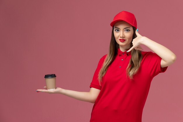 Vue de face femme courrier en uniforme rouge et cape tenant la tasse de café de livraison sur le travail uniforme de prestation de services de fond rose