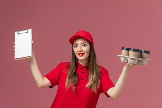 Photo gratuite vue de face femme courrier en uniforme rouge tenant le bloc-notes de tasses de café de livraison souriant sur fond rose service de livraison de l'entreprise de travail uniforme