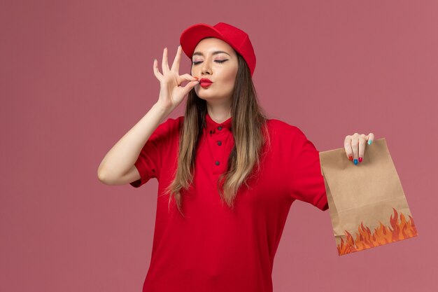 Vue de face femme courrier en uniforme rouge tenant le paquet alimentaire papier montrant savoureux signe sur fond rose service de livraison de travail entreprise uniforme