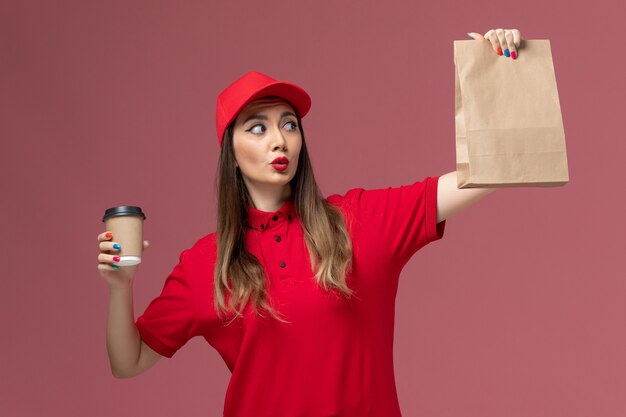 Vue de face femme courrier en uniforme rouge tenant la tasse de café de livraison et le paquet de nourriture sur le travail uniforme de travailleur de livraison de services de fond rose