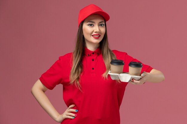 Vue de face femme courrier en uniforme rouge tenant des tasses de café de livraison brun souriant sur le travail uniforme de livraison de services de fond rose