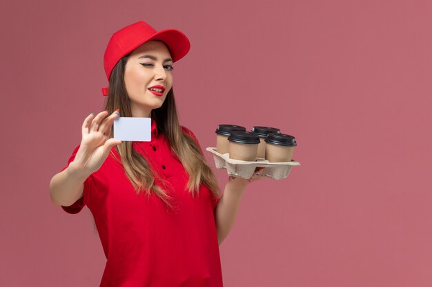 Vue de face femme courrier en uniforme rouge tenant des tasses de café de livraison et carte clignotant sur fond rose service entreprise de livraison de travail uniforme