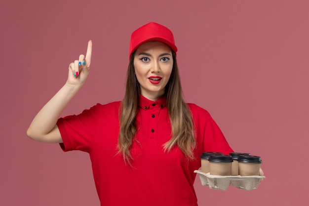 Vue de face femme courrier en uniforme rouge tenant des tasses de café de livraison en levant le doigt sur l'uniforme de livraison de services d'emploi de fond rose