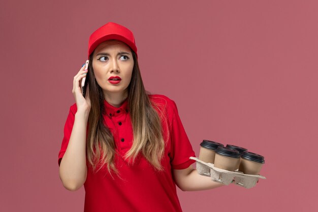 Vue de face femme courrier en uniforme rouge tenant des tasses de café de livraison parler au téléphone sur l'uniforme de prestation de services de fond rose