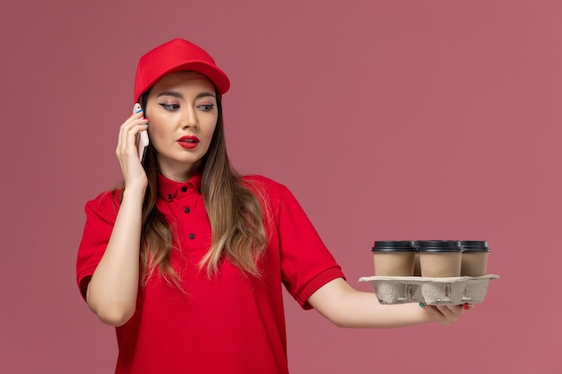 Vue de face femme courrier en uniforme rouge tenant des tasses à café et parler au téléphone sur l'uniforme de livraison de service de bureau rose