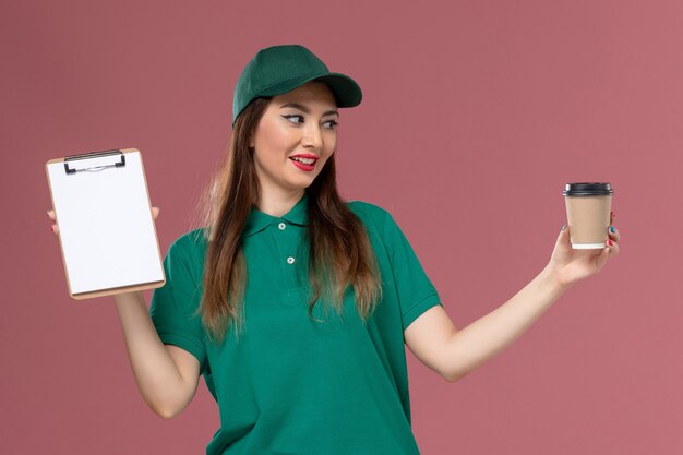 Vue de face femme courrier en uniforme vert et cape tenant la tasse de café de livraison et le bloc-notes sur le mur rose service travail de livraison uniforme de travail entreprise