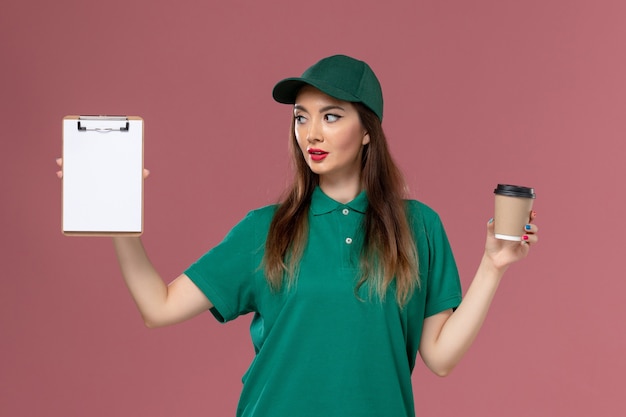 Vue de face femme courrier en uniforme vert et cape tenant la tasse de café de livraison et le bloc-notes sur le service de bureau rose travail de livraison uniforme travail travailleur féminin entreprise