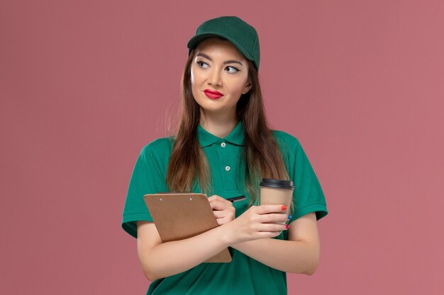 Vue de face femme courrier en uniforme vert et cape tenant la tasse de café de livraison et le bloc-notes stylo écrit sur le mur rose entreprise service emploi livraison uniforme