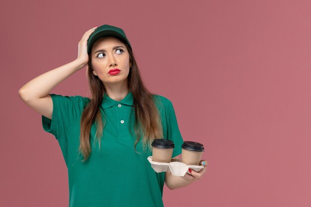 Vue de face femme courrier en uniforme vert et cape tenant des tasses de café de livraison en pensant sur le mur rose entreprise service emploi livraison uniforme